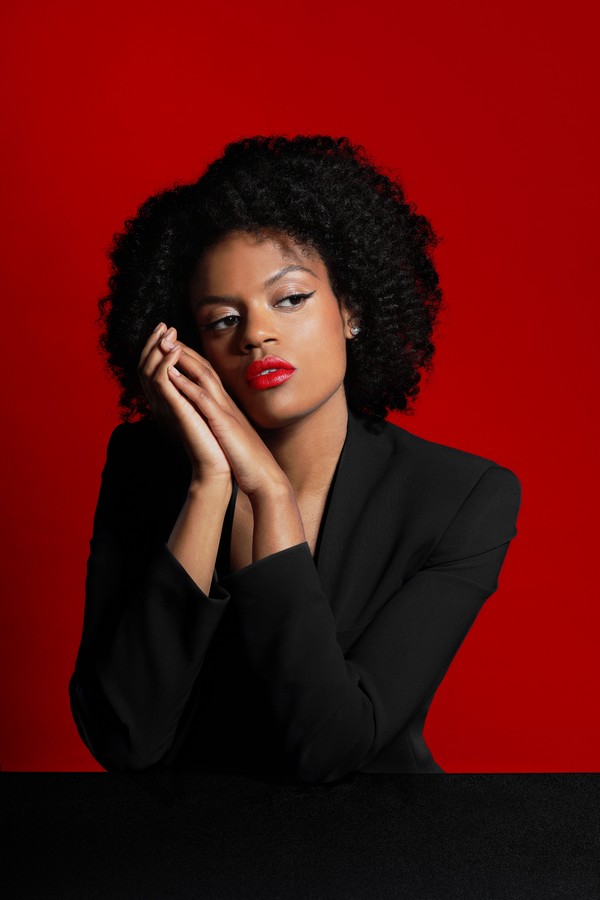 Portrait studio d'Axelle Saint-Cirel sur fond rouge vif, portant un blazer noir, maquillage élégant avec rouge à lèvres rouge, mise en scène artistique
